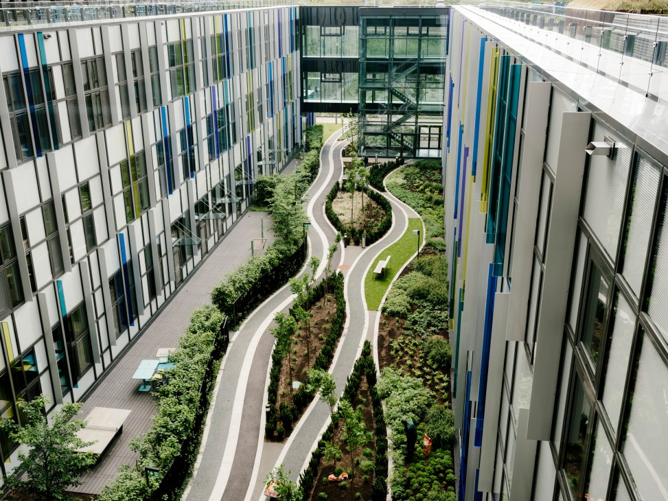 Aerial view of a landscaped courtyard garden with winding pathways, trees, and green space surrounded by modern hospital buildings with blue and white facade.