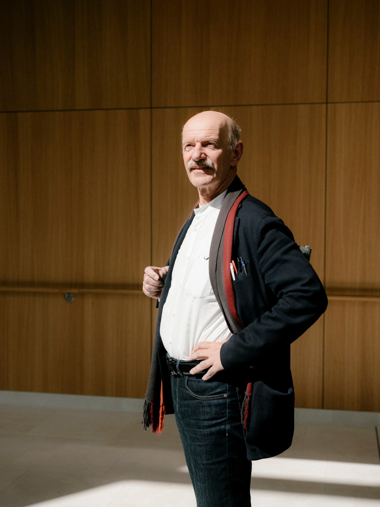 Architect Albert Wimmer stands against a wooden wall, wearing a black blazer with a red scarf over a white shirt.