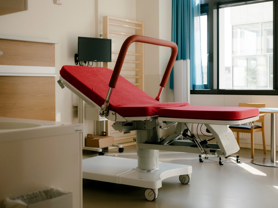 Red examination chair with curved armrests in a bright clinical room with natural light from windows.