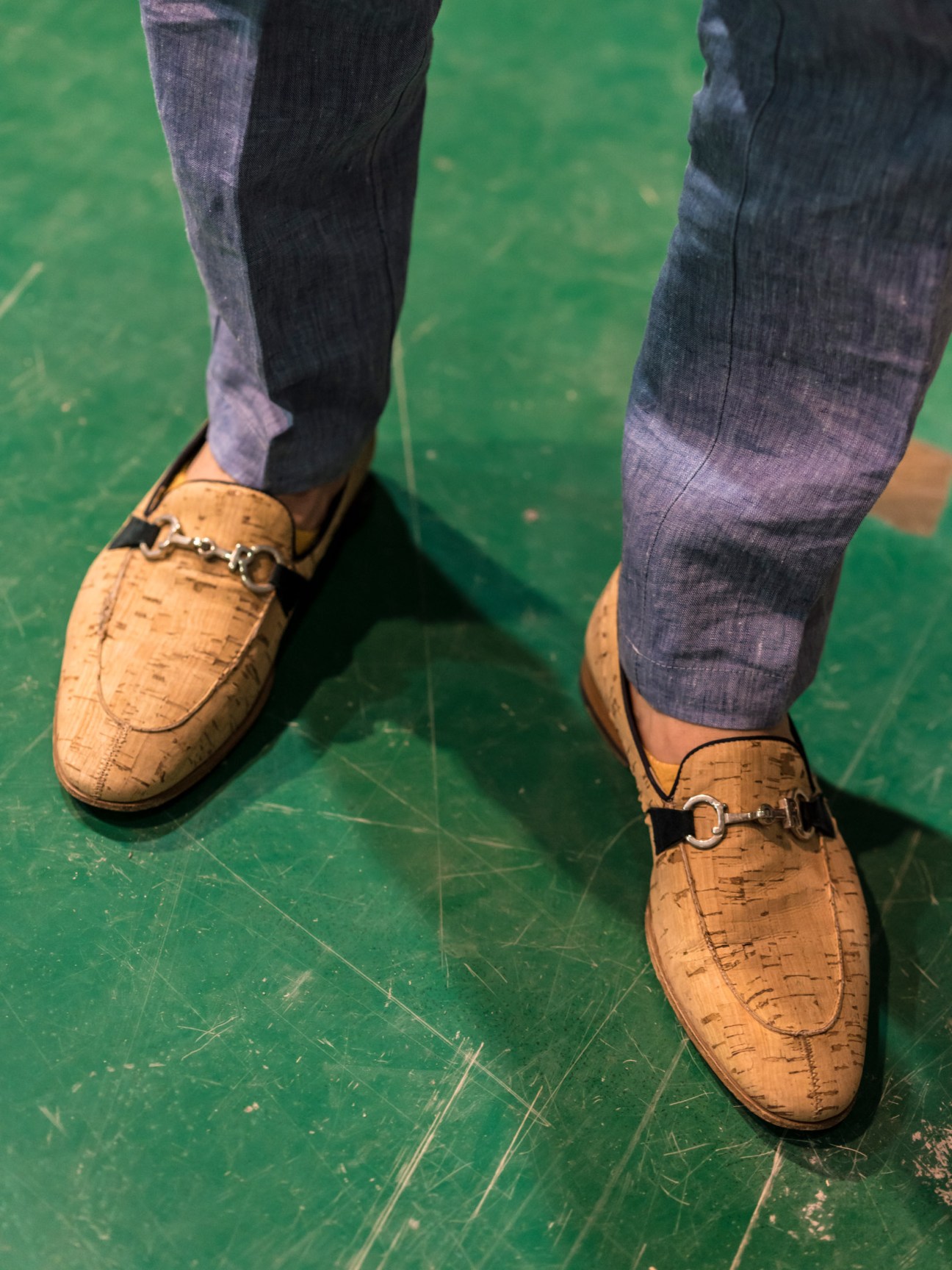 Cork-textured loafers with metal hardware worn with grey socks on a green floor.