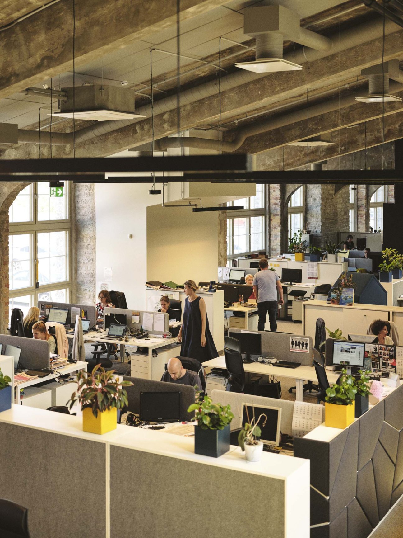 Open-plan office with workstations arranged around the perimeter of a large industrial space with concrete columns and pitched glass roof.