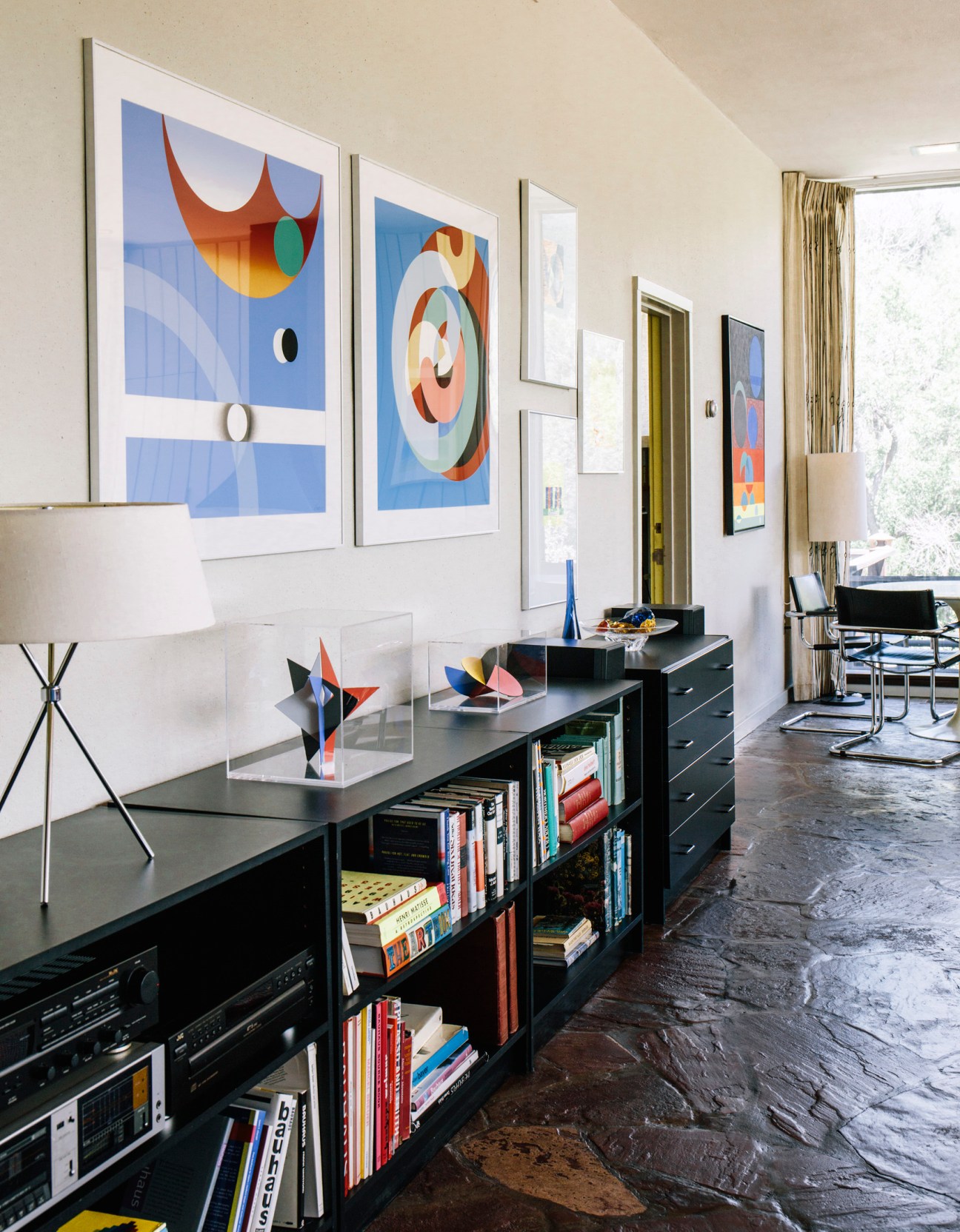 Modern office with black desk, books, colorful abstract artwork on white walls, and mountain views through windows.
