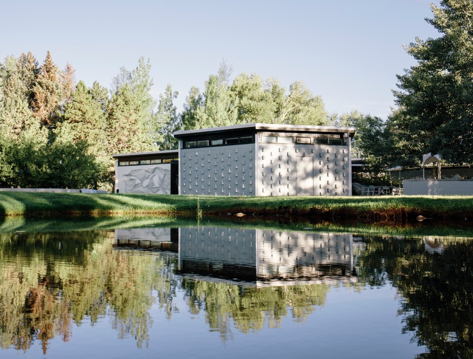 Modern white building with geometric perforated facade reflected in calm water, surrounded by trees.