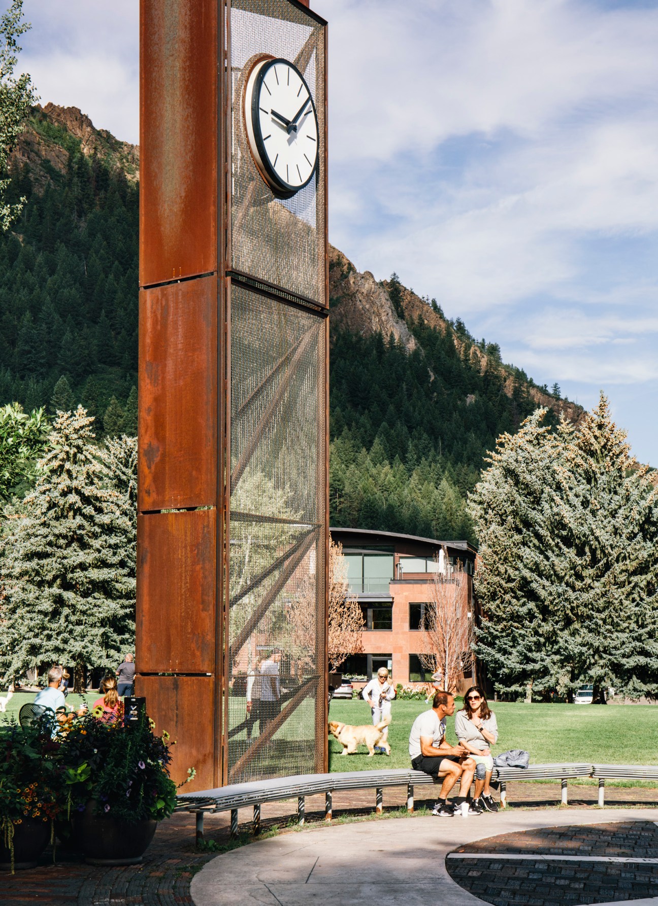 Modern architectural clock tower with rusted steel and mesh panels set against Rocky Mountain backdrop.