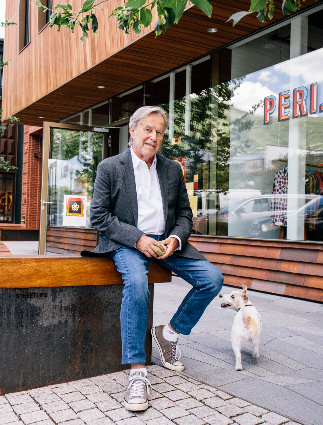 Artist Dick Carter sitting on a bench outside a storefront in Aspen, wearing a blazer and jeans with a small white dog nearby.