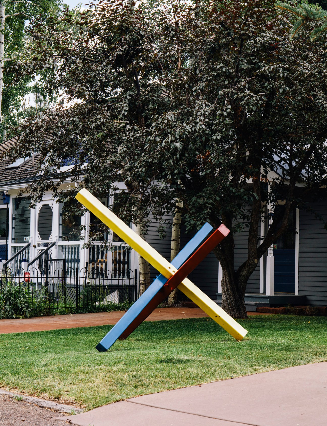 Colorful geometric sculpture with yellow, blue, and red beams arranged in an X-shape on a lawn in front of a historic house and large tree.