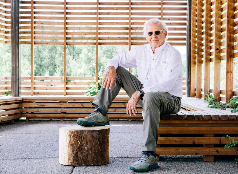 Architect Harry Teague sits on a wooden bench wearing sunglasses and white shirt in a modernist structure with wooden lattice screening.
