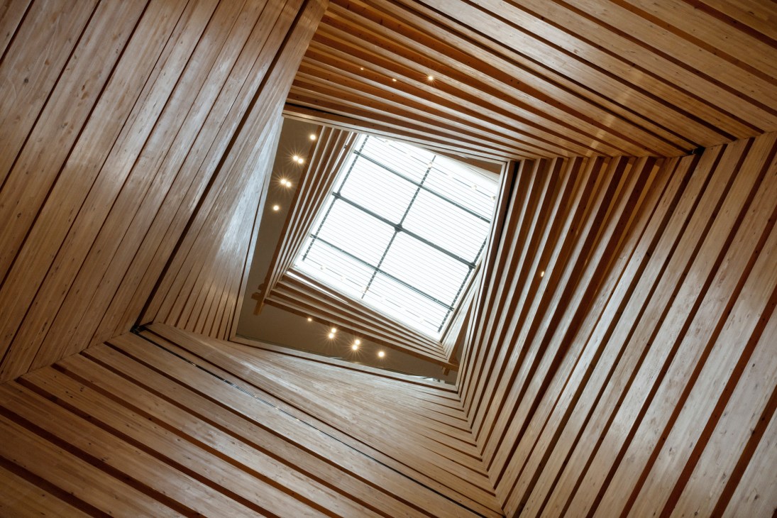 Upward view of a square skylight framed by concentric undulating timber beams in warm pine tones with recessed lighting.
