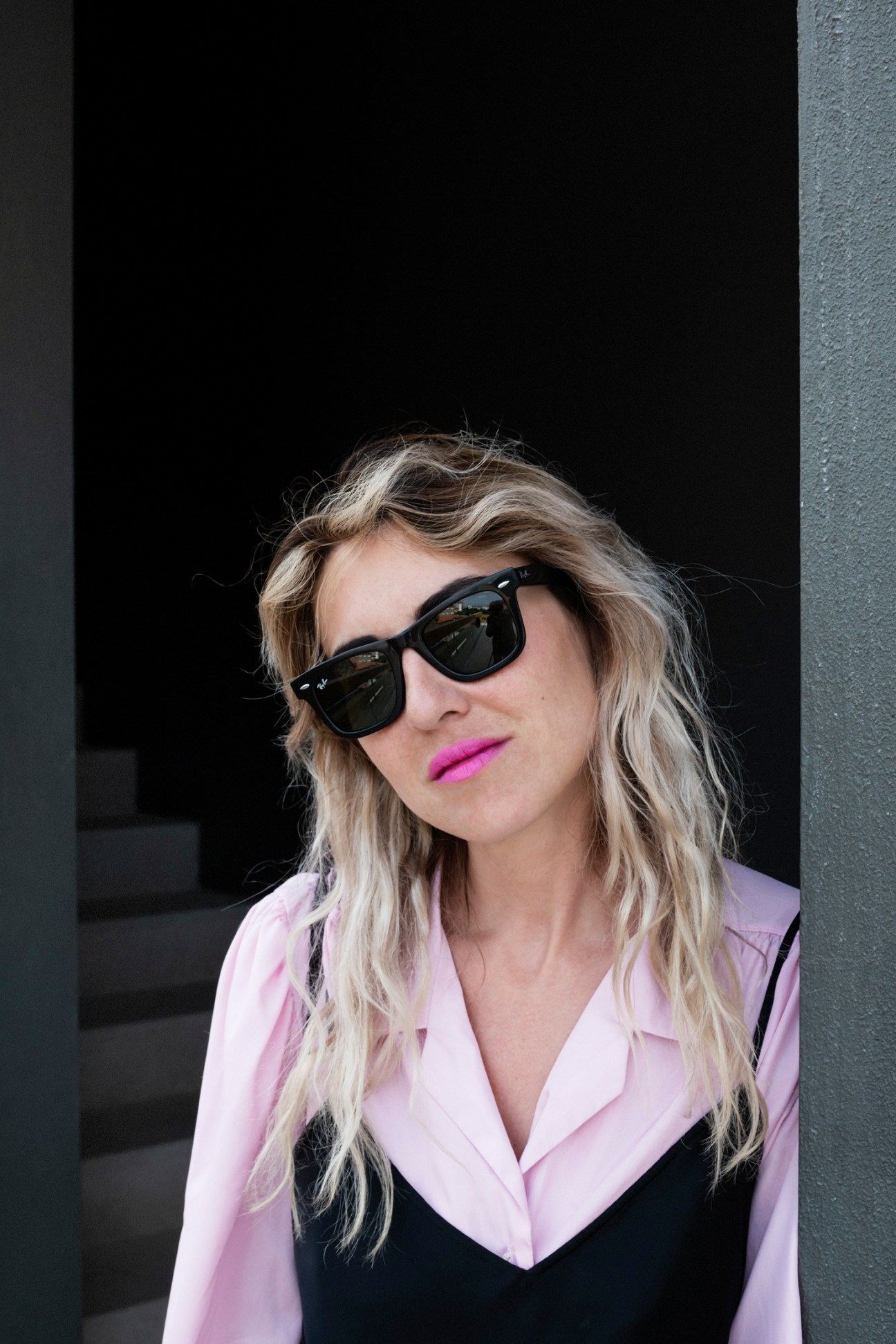 Idil Tabanca wearing black sunglasses and pink blazer standing against dark background.