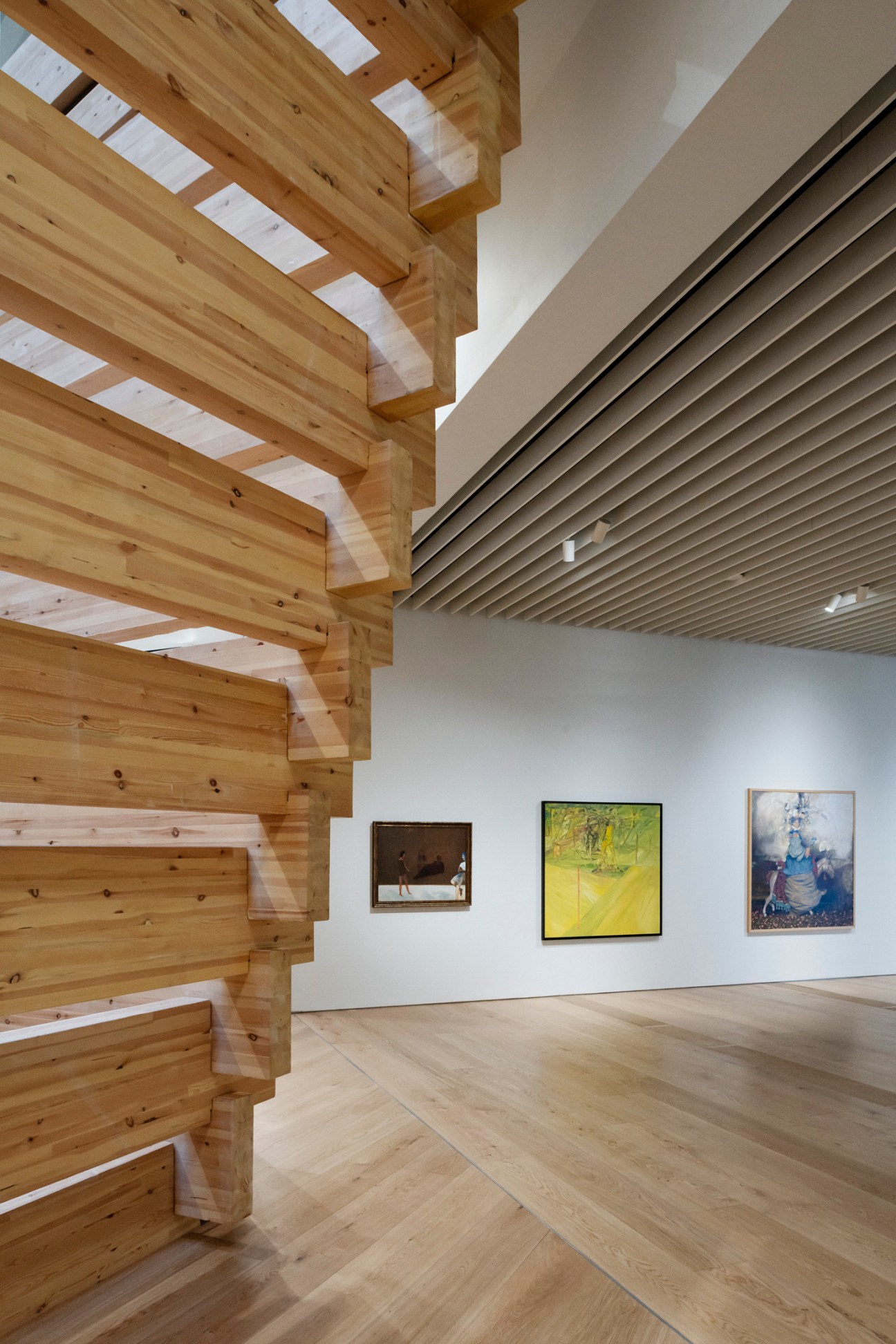Museum gallery interior with wooden structural beams filtering light from above onto white walls displaying framed artworks.