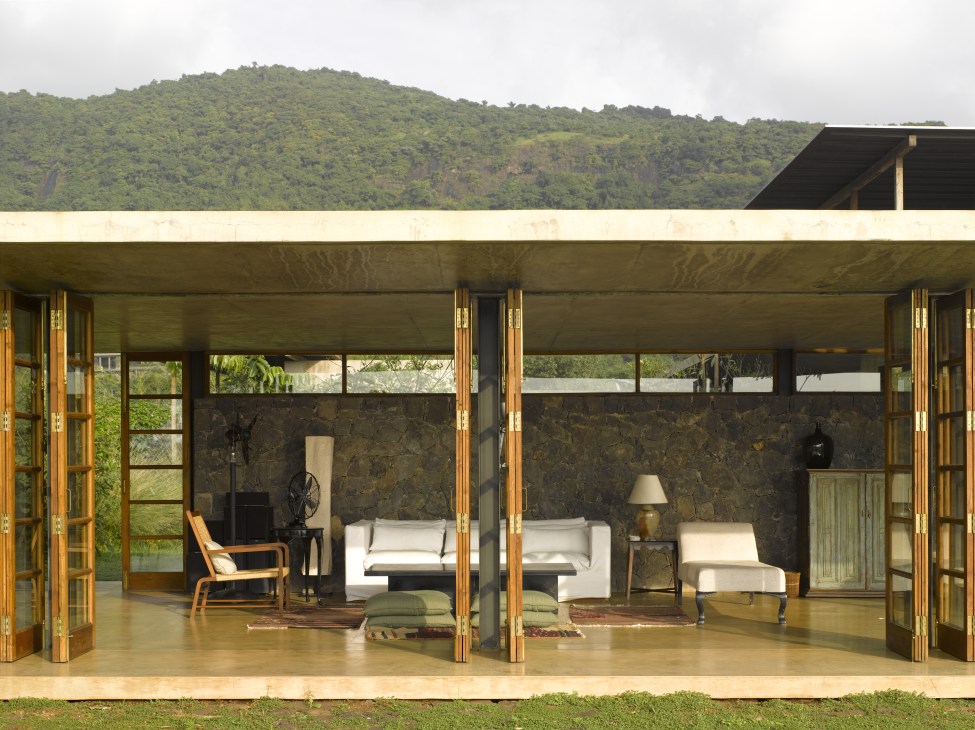 Modern living room with wooden doors opening onto forested landscape, featuring stone walls and contemporary furnishings.