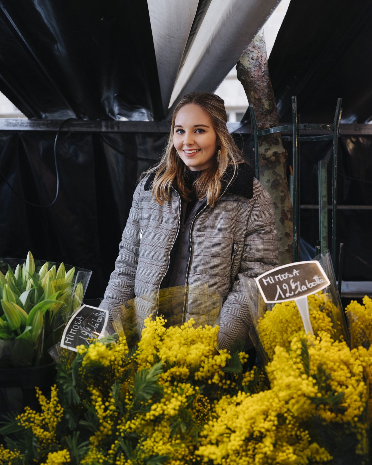 Young woman in plaid jacket standing behind yellow mimosa flowers at a market stall.
