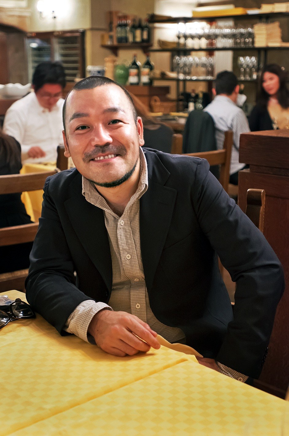 Kentaro Kobayashi smiling at a table inside La Bisboccia restaurant with diners and shelved bottles visible in the background.