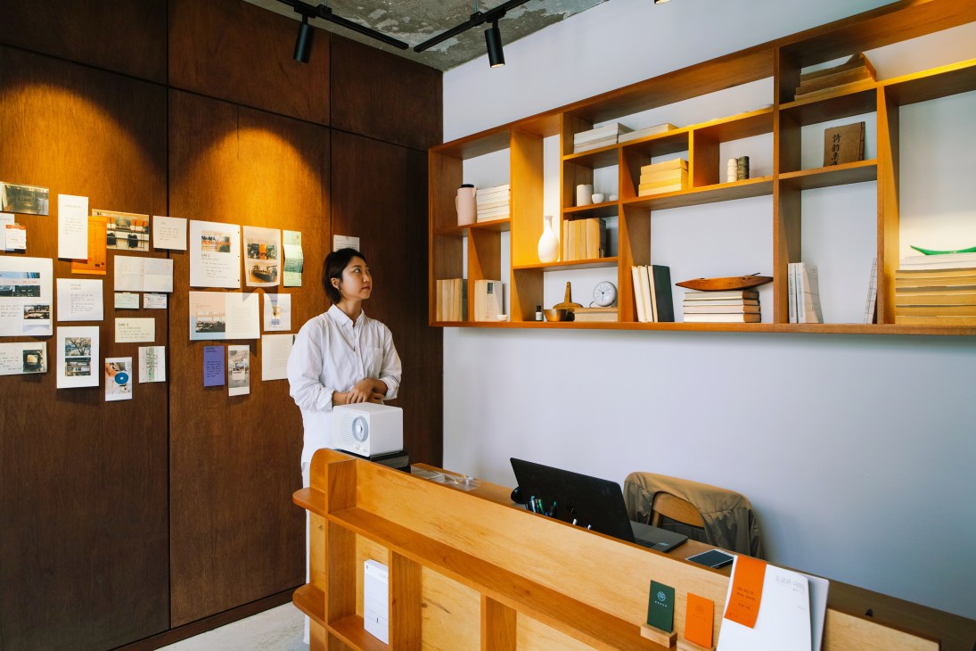 Woman in white shirt standing behind wooden counter in bookshop with wooden shelving and displayed books behind her.