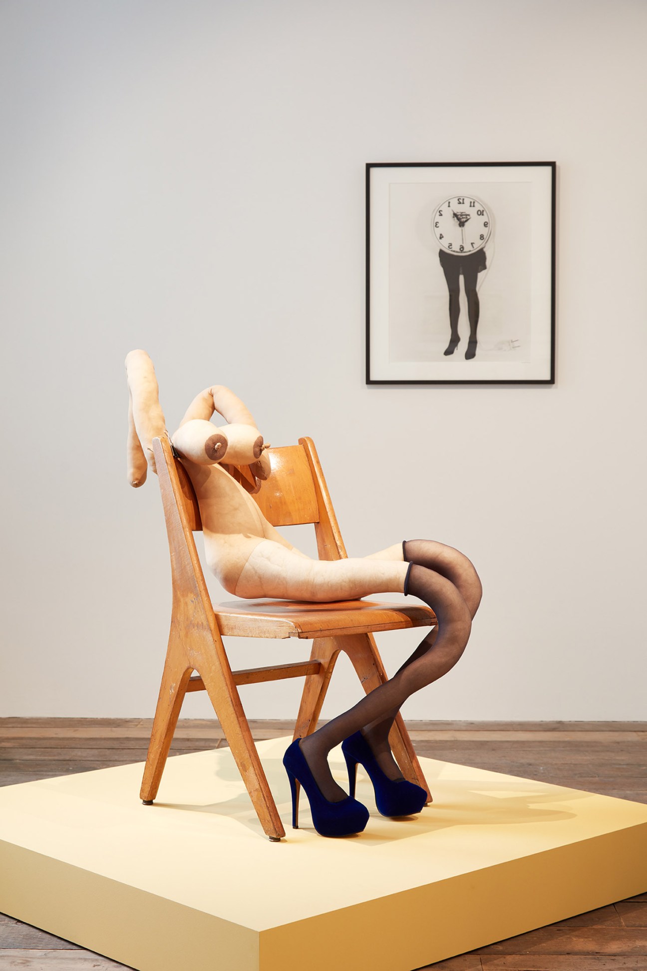 Surreal sculpture of a wooden chair with a pale mannequin torso and dark legs wearing heels, displayed in a gallery space with a framed artwork above.