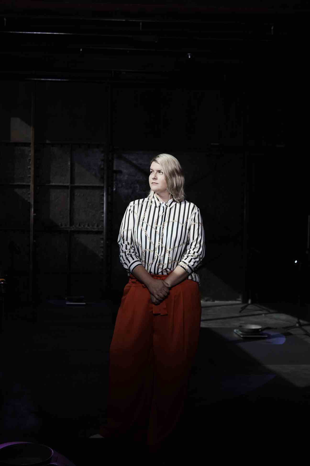 Sarah McCrory, director of Goldsmiths Centre for Contemporary Art, standing in the gallery space wearing a striped shirt and red trousers.