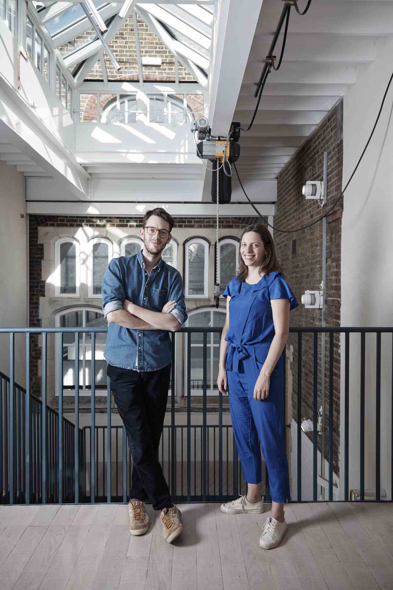 Adam Willis and Paloma Strelitz of Assemble stand in a bright gallery space with skylights and exposed brick.