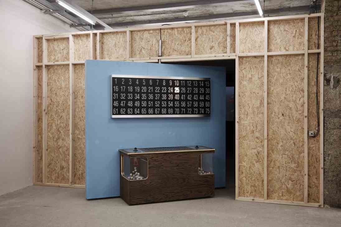 Installation by Mika Rottenberg featuring wooden partitions, a blue wall, numbered display board, and dark wooden table with objects.