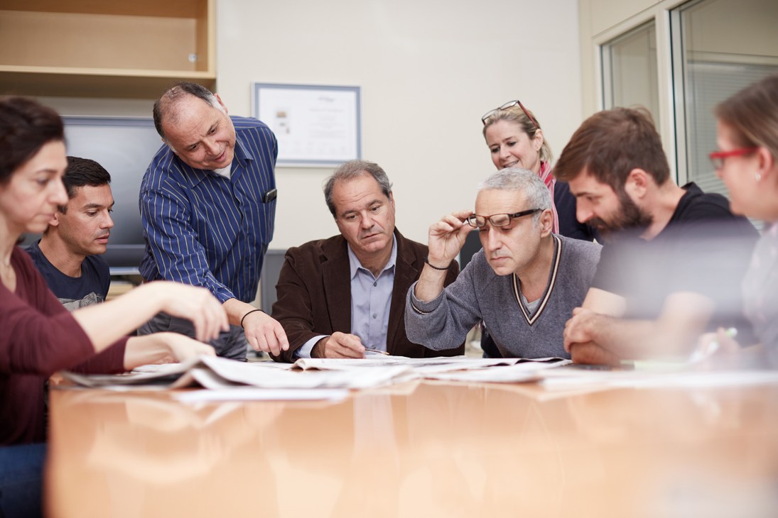 Kathimerini English edition team members gathered around a table reviewing documents in an office setting.