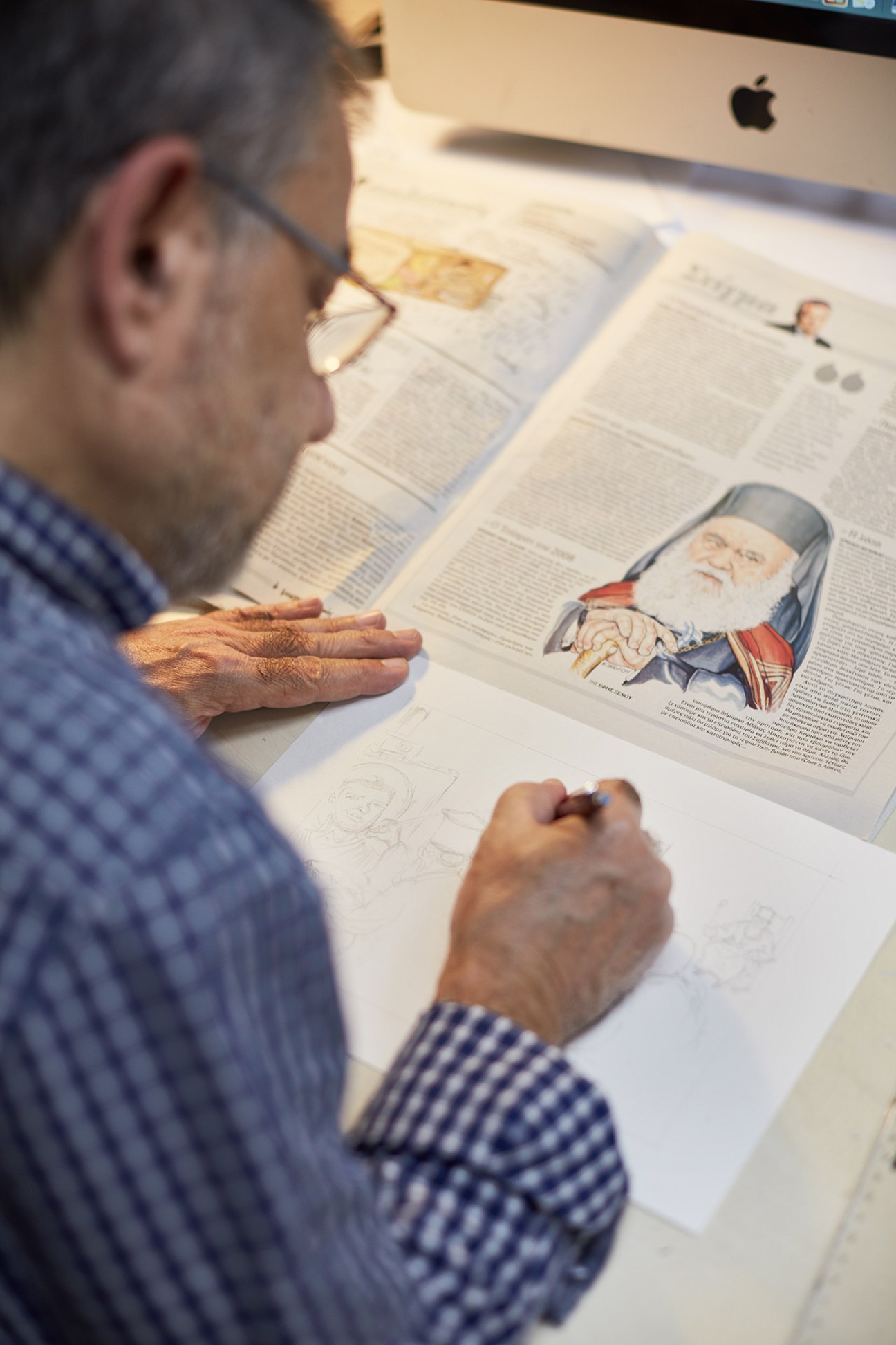Cartoonist Elias Makris sketches at his desk at Kathimerini newspaper with a completed cartoon visible.