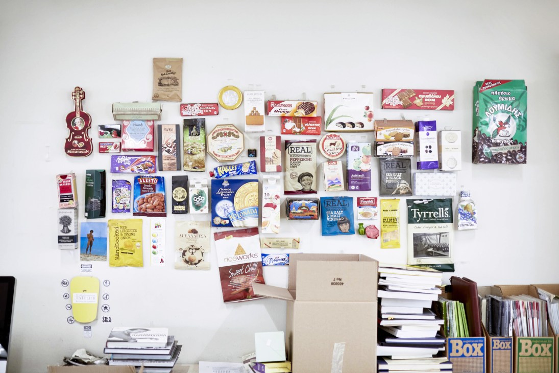 Flat lay of Greek food and drink products arranged on a white wall, including packaging, boxes, bottles, and a decorative guitar.
