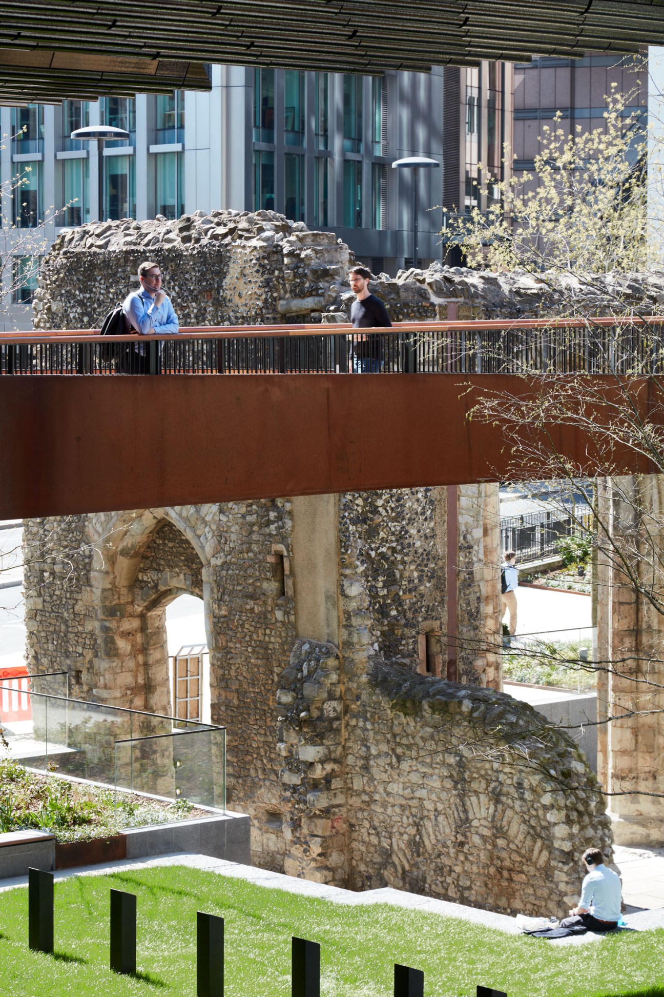 Two people walk on an elevated wooden walkway with Roman stone ruins and modern glass buildings visible in the background.