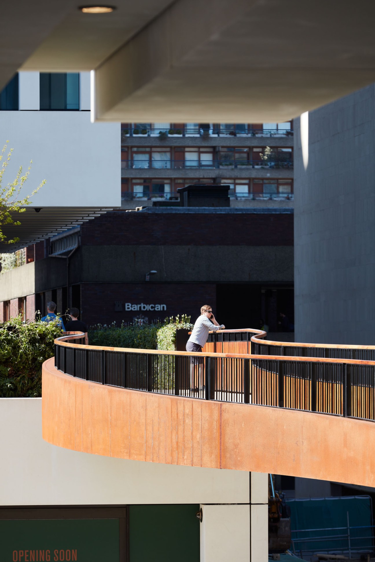 Person leaning on railing of elevated wooden walkway with Barbican Centre brutalist building visible below