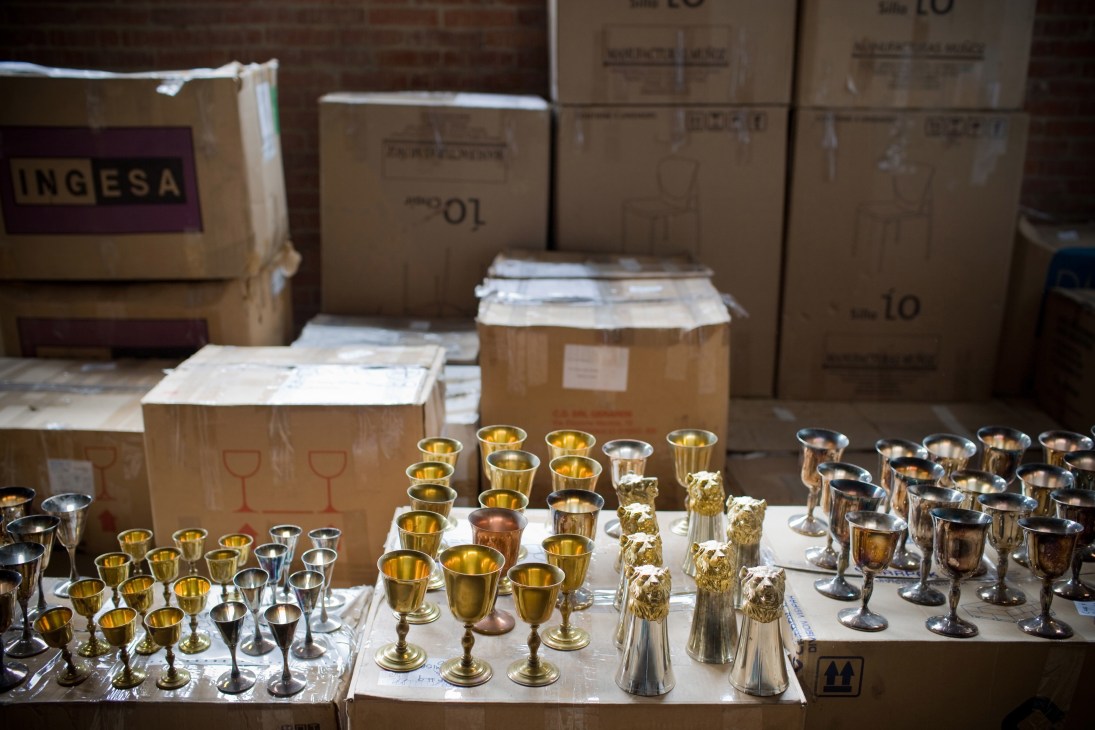 Gold and silver goblets and chalices displayed on white platforms in front of cardboard storage boxes in a warehouse.