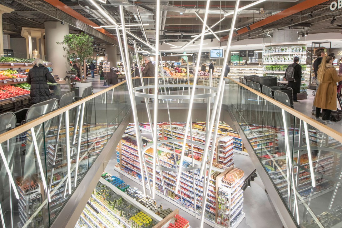 Interior view of Zurheide supermarket showing two floors with illuminated product displays and customers shopping.