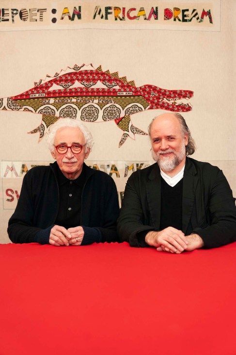 Two men seated at a red table beneath colorful patterned artwork featuring an African animal motif.