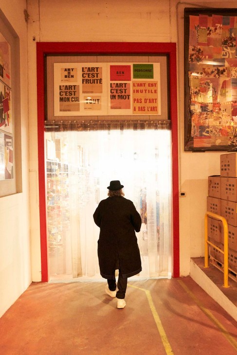 Man in black coat and hat walks toward a large red-framed window with French text artwork above it in a factory setting.