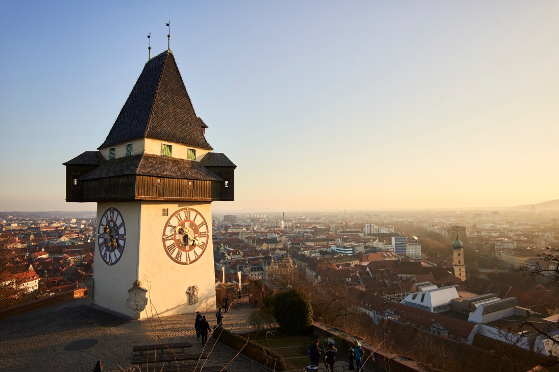 Clock tower with wooden gallery and pointed roof overlooking red-roofed cityscape at sunset