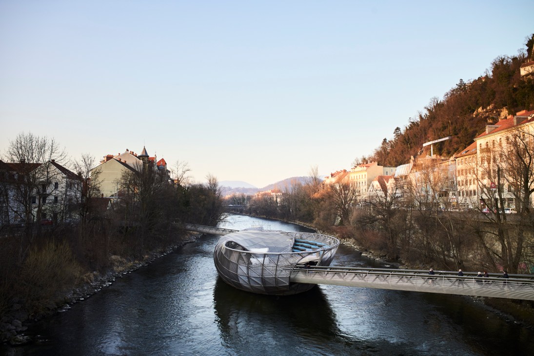 Modern circular steel bridge structure spanning river with historic colorful buildings on hillsides at dusk