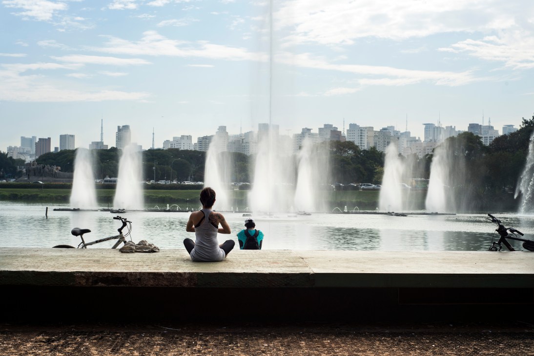 Woman and child sitting on sandy shore of Ibirapuera Park watching fountains on lake with São Paulo skyline beyond.