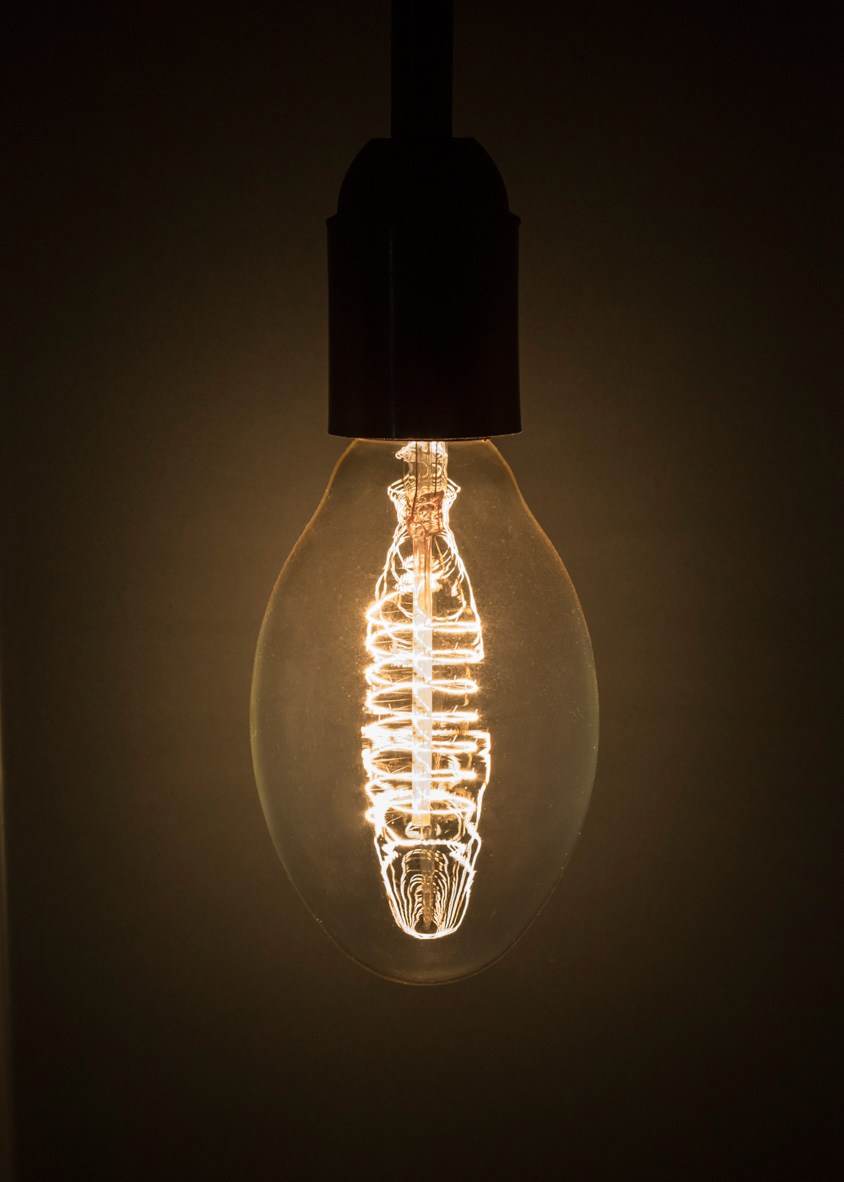 Illuminated incandescent bulb with intricate tungsten filament glowing warm amber against dark background.