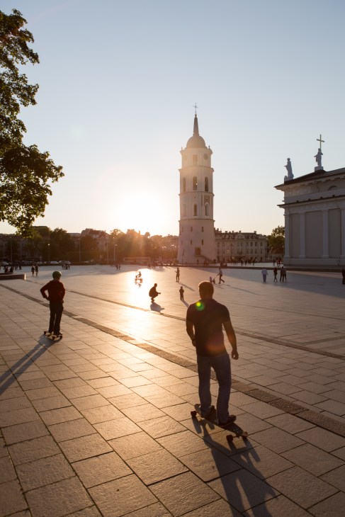 People skateboarding and walking in Cathedral Square at sunset with a pink bell tower and cathedral in the background.