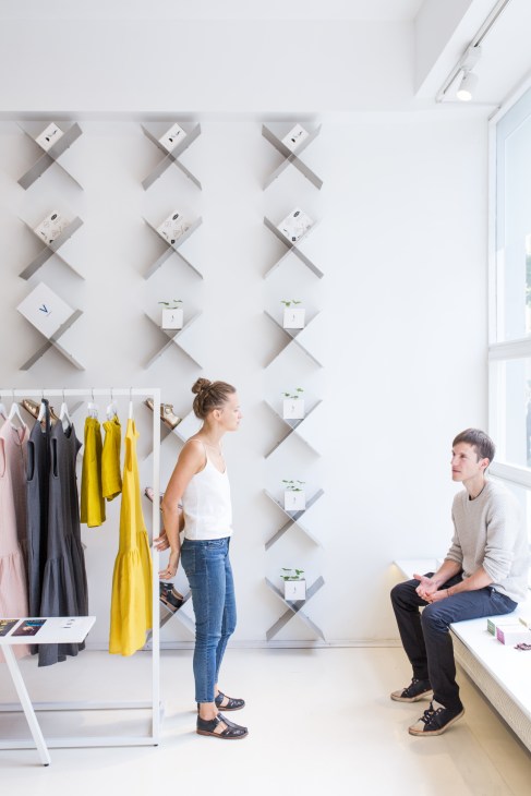Two people stand in a minimalist retail space with X-shaped shelving units displaying products and plants on a white wall.