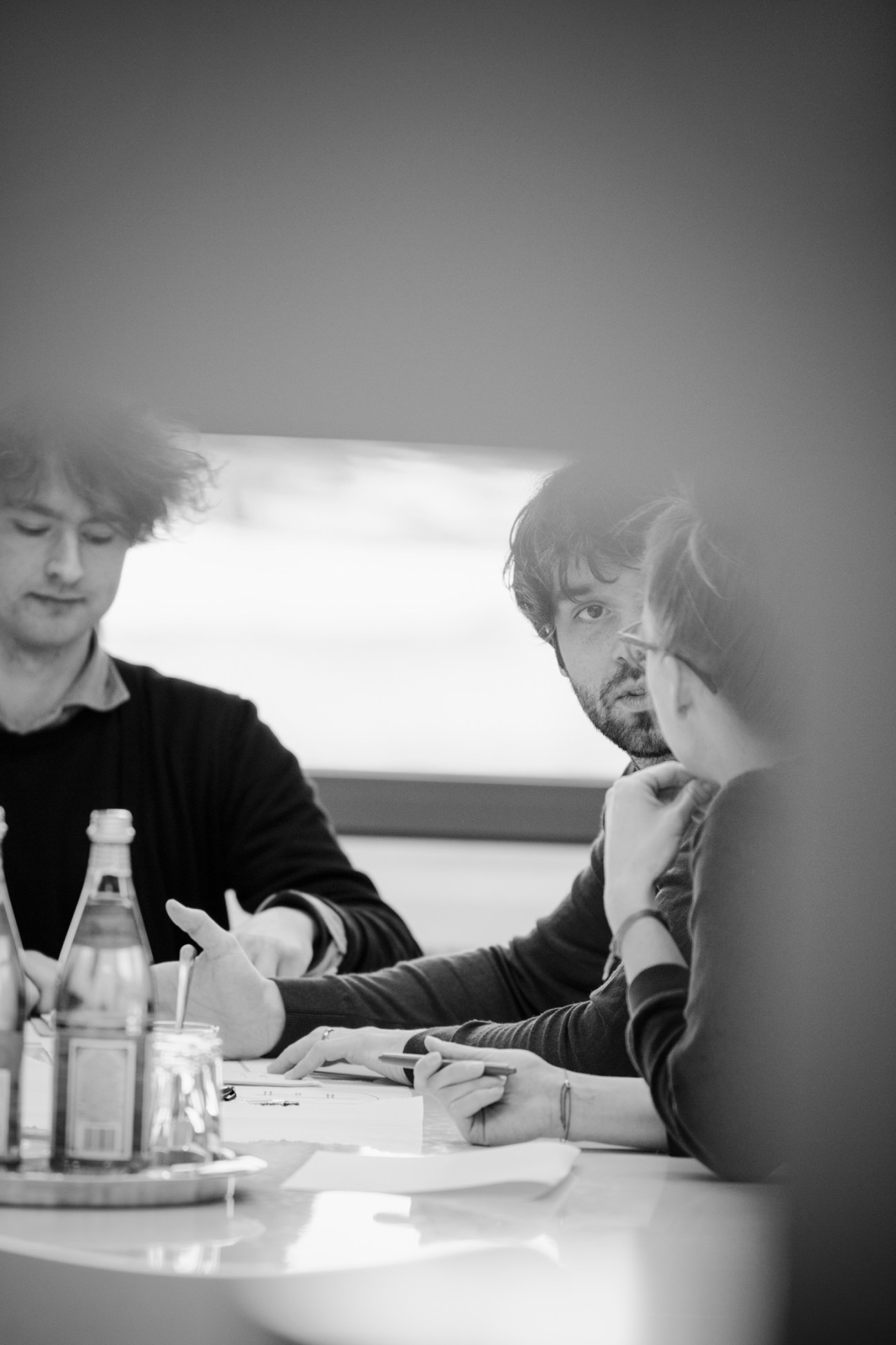 Two men in conversation at a table with water bottles and papers in black and white