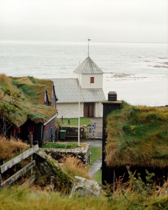 A white Faroese church with turf-covered hillsides, overlooking the ocean under cloudy skies.