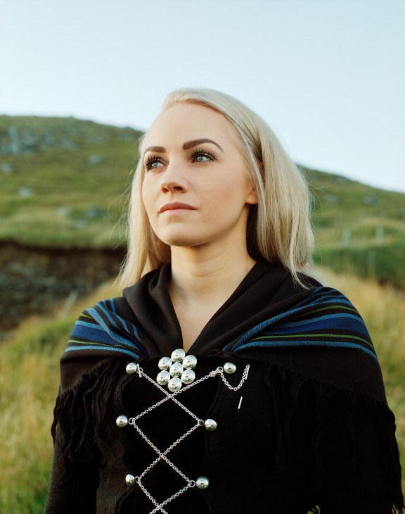 Woman in traditional Faroese dress with black wool tunic and silver chain brooch against green hillside.