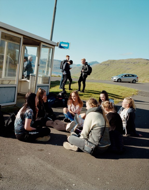 A group of people sit on a road beside a bus shelter with green mountains visible in the Faroe Islands landscape.