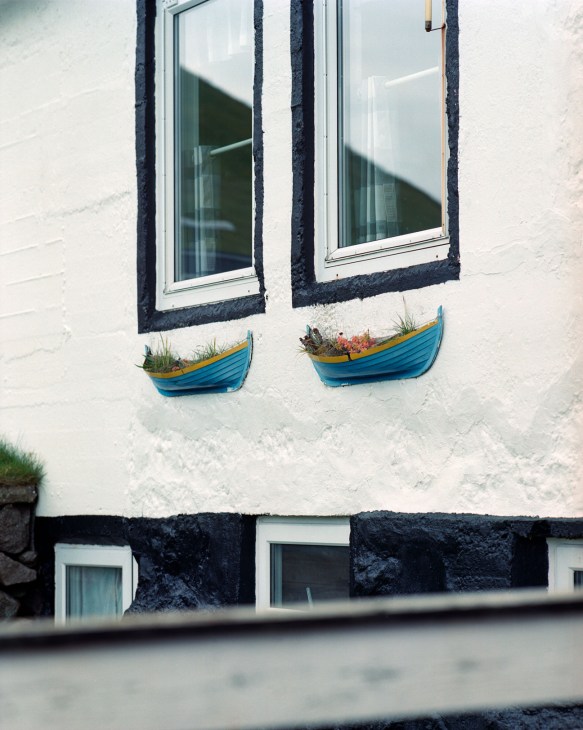 White-painted Faroese house with black trim, two windows with dark frames, and blue decorative boat planters beneath each window.