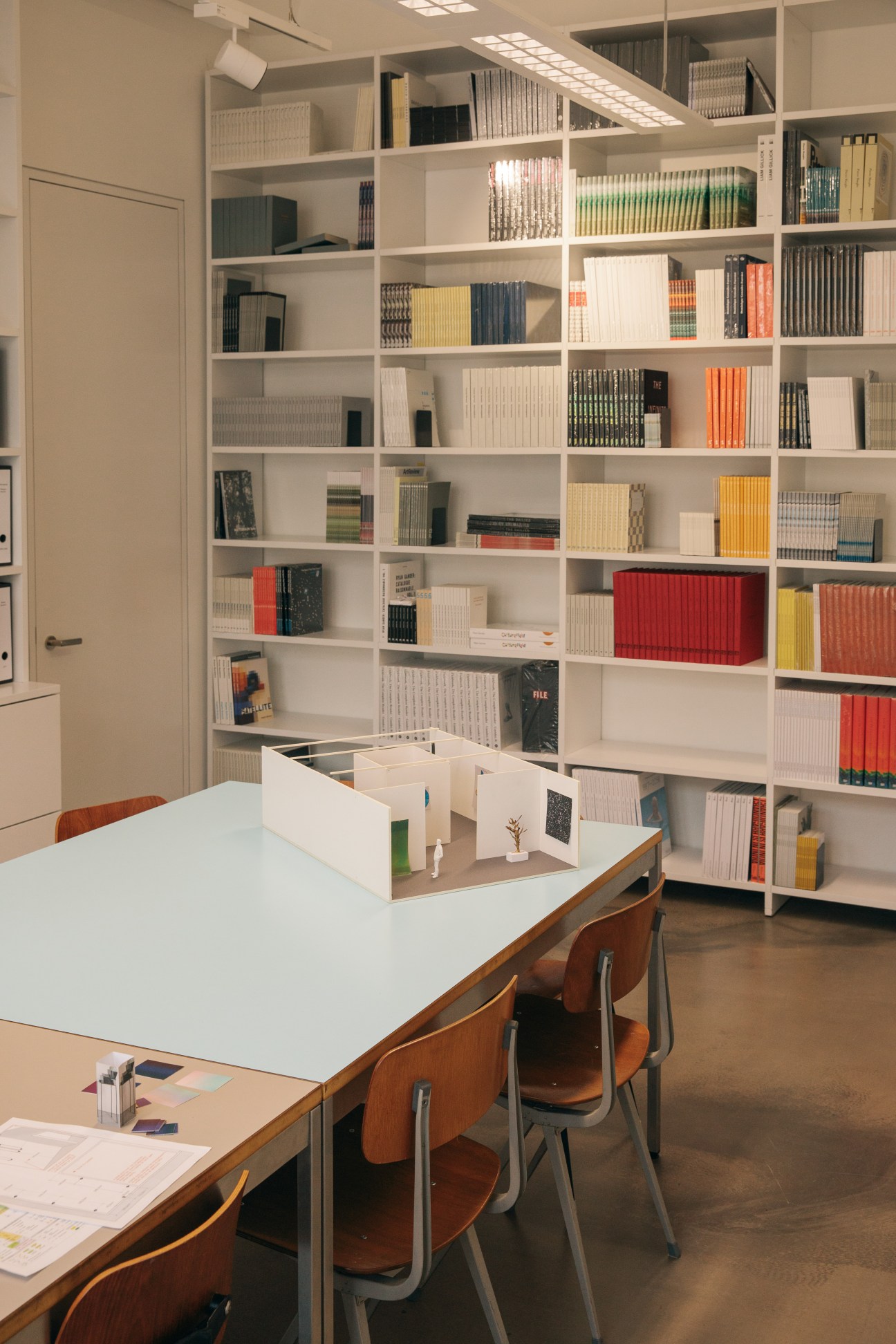 White shelving unit filled with art catalogues and archives behind a work table with a scale model of an exhibition booth.