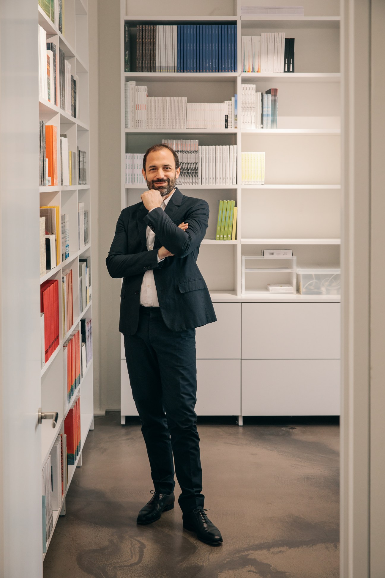 Florian Wojnar stands posing in front of white shelving filled with books and art catalogues in a model-making room.