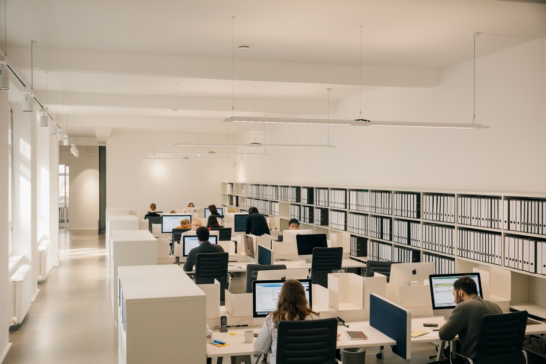 Gallery employees working at desks with computers in a bright, minimalist office space with filing cabinets and pendant lighting.