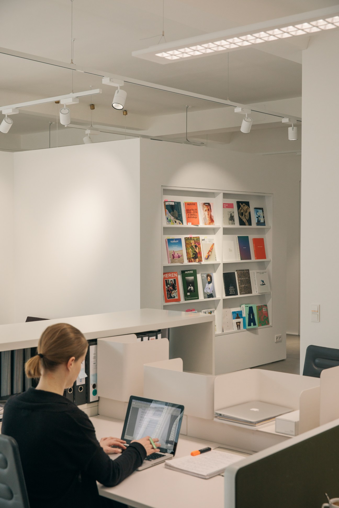 Gallery staff member working at desk in minimalist white office with shelves of art catalogues.