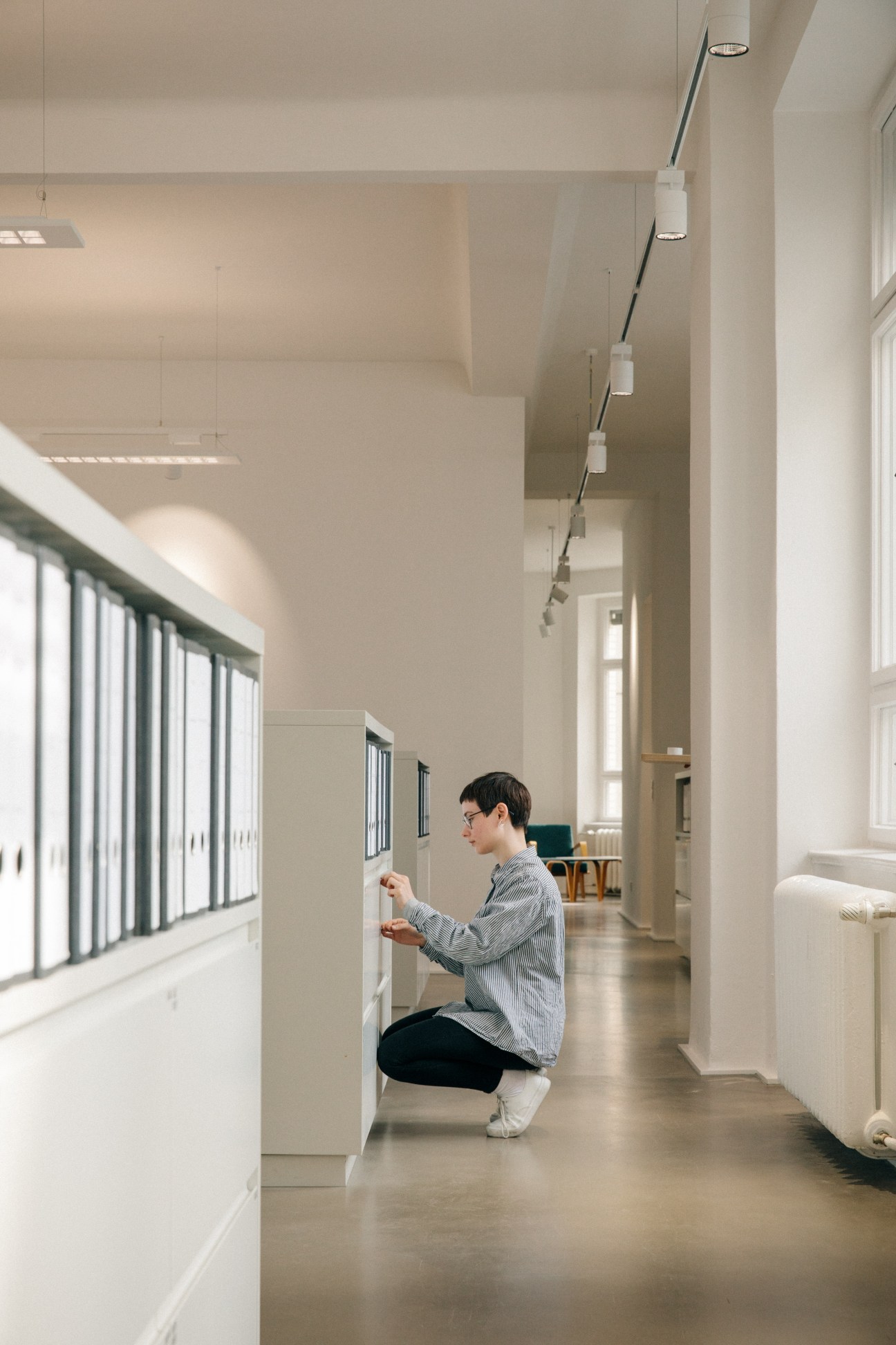 Gallery staff member organizing files in a bright, minimalist office corridor with filing cabinets.