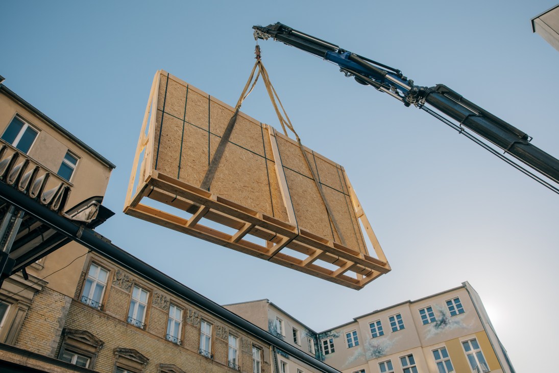 A wooden art installation with metal framework suspended by ropes from a blue crane over a Berlin courtyard.