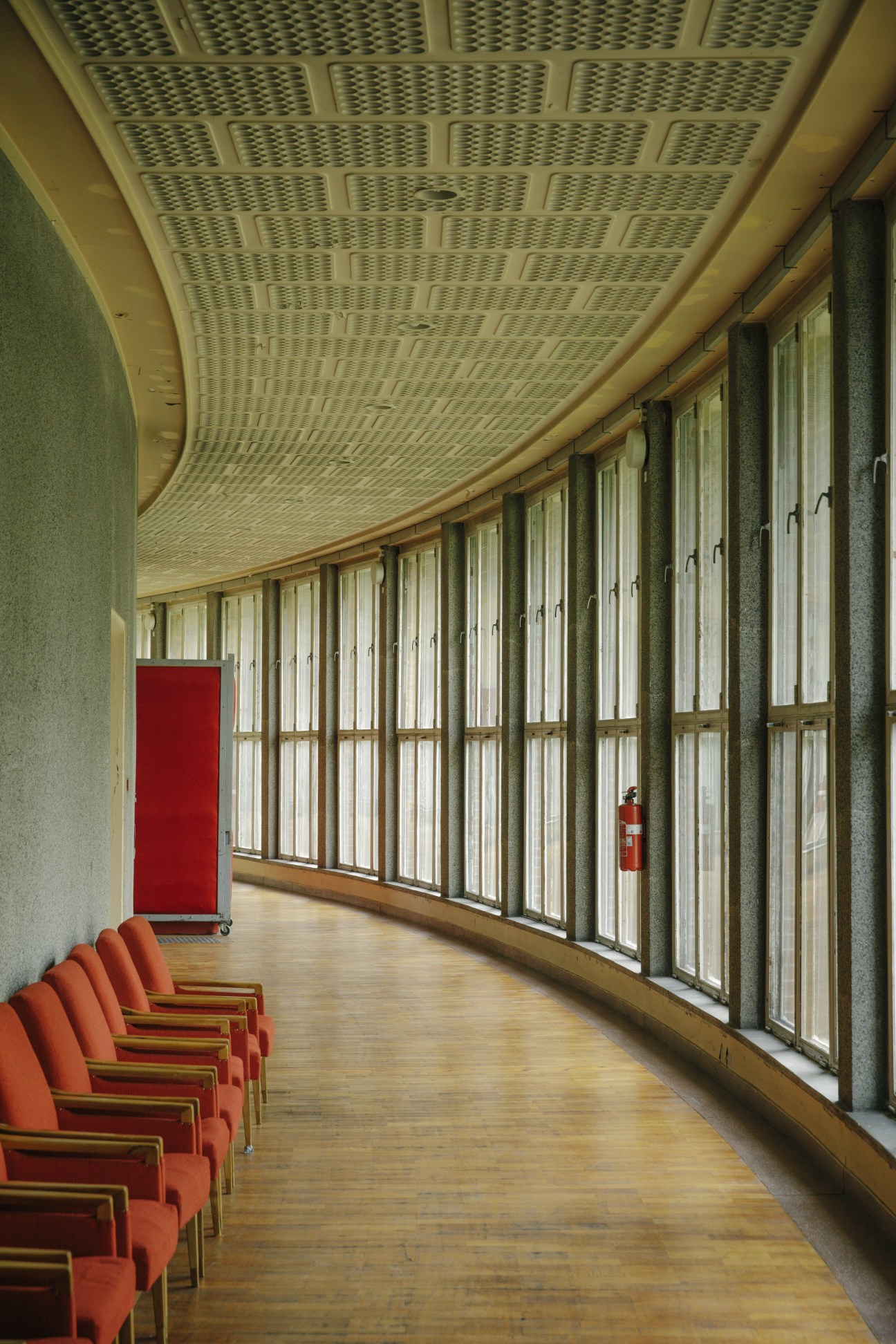 Modernist corridor in Block B with perforated ceiling, floor-to-ceiling windows, orange seating, and red fire extinguisher.