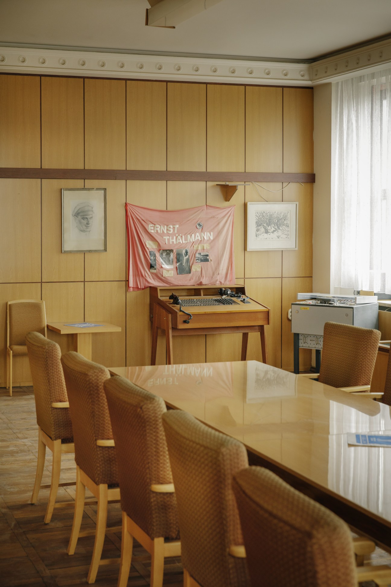 A mid-century modern directors' office with wood-paneled walls, period furniture, and a pink banner reading 'Ernst Thalmann' hanging above vintage broadcasting equipment.